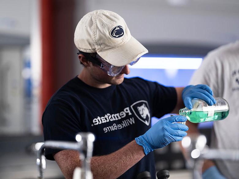 male student wearing a Penn State cap, goggles, and laytex gloves pouring a bright green liquid into a beaker