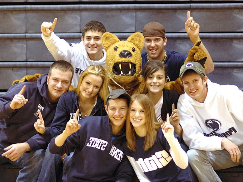 Students sitting on bleachers and gathered around the Nittany Lion mascot, facing forward and holding up their index fingers in the “we're number one” sign